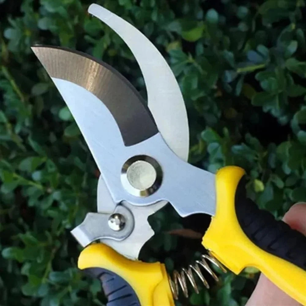 A close-up of a yellow and black bypass pruning shears with their stainless steel blades wide open. The shears are held in a hand, with a background of green leaves