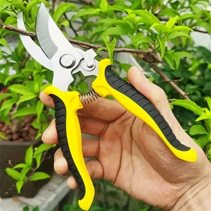 A hand holds a pair of yellow and black shears for gardening with stainless steel blades. The shears are positioned to prune a small branch on a leafy green plant.