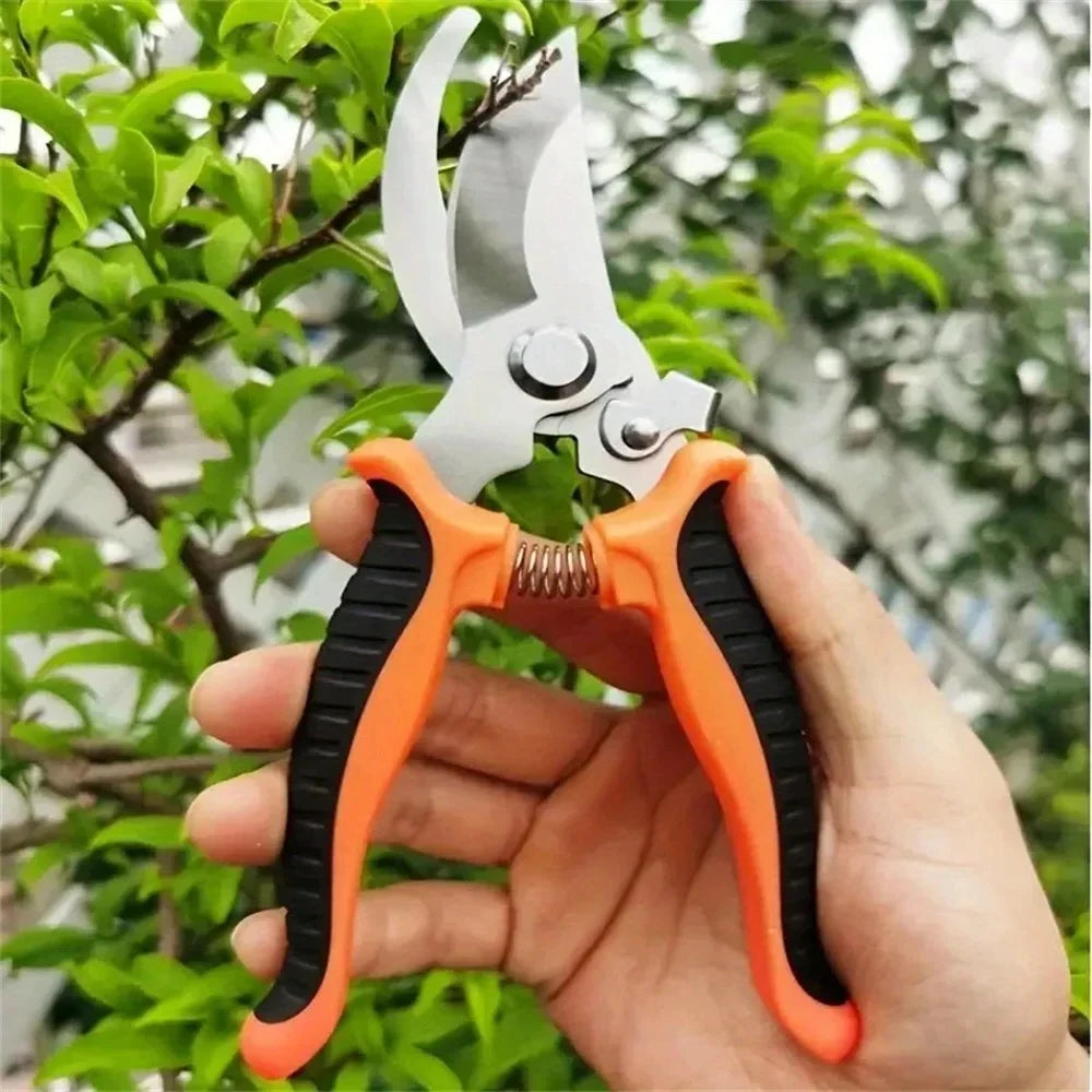 A hand holds a orange and black shears garden tool with stainless steel blades. The shears are open, ready to prune a leafy green plant in the background.